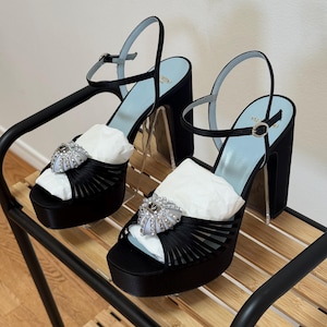 May include: A pair of black satin platform sandals featuring a high block heel, an adjustable ankle strap with a silver buckle, and a light blue insole. The front of each shoe is adorned with a decorative silver-toned heart-shaped embellishment.