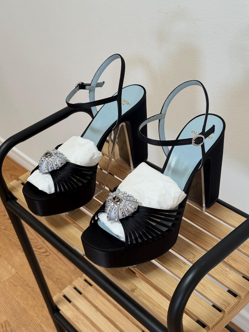 May include: A pair of black satin platform sandals featuring a high block heel, an adjustable ankle strap with a silver buckle, and a light blue insole. The front of each shoe is adorned with a decorative silver-toned heart-shaped embellishment.