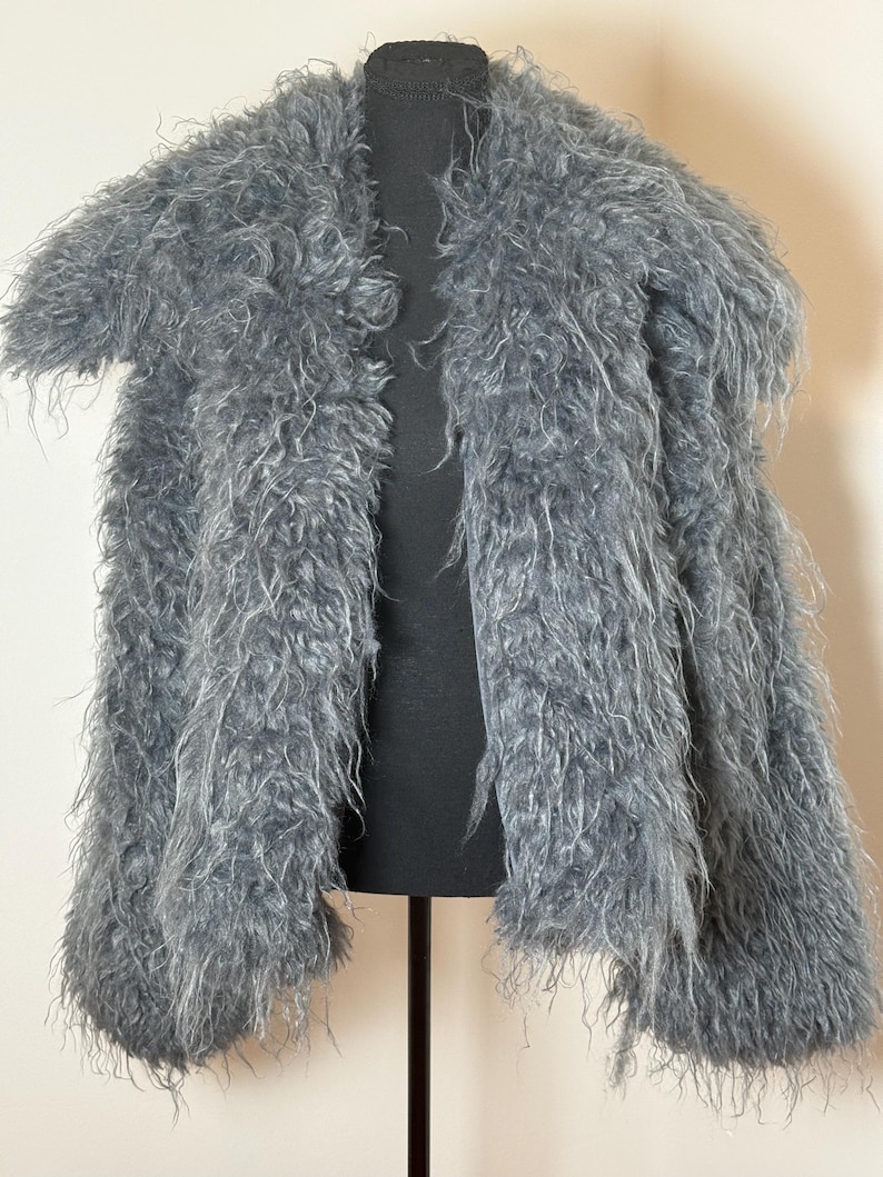 May include: A gray, shaggy faux fur coat displayed on a black mannequin. The coat has a collarless design and short sleeves. The fur appears soft and has a textured appearance, with varying shades of gray.