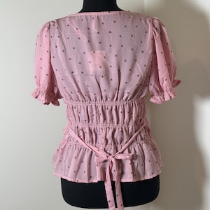 May include: A light pink blouse with short, ruffled sleeves and a cinched waist. The blouse features a delicate pattern of small, dark brown floral motifs. A matching pink tie belt cinches the waist, and the back is visible.