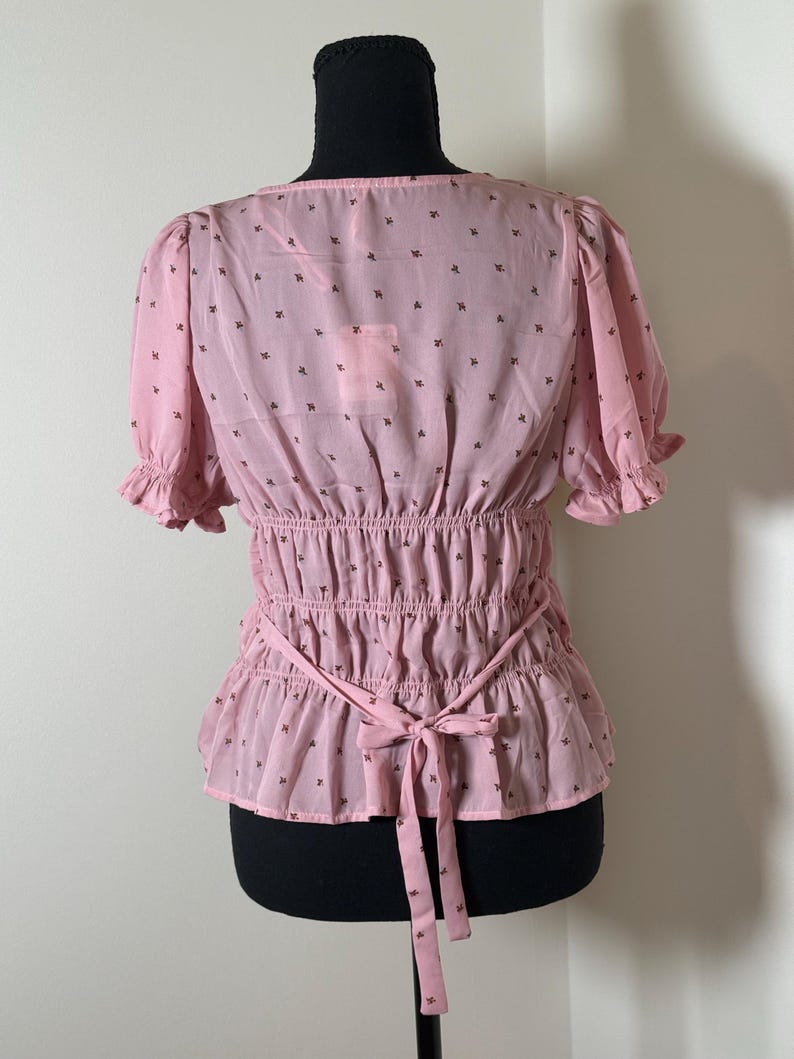 May include: A light pink blouse with short, ruffled sleeves and a cinched waist. The blouse features a delicate pattern of small, dark brown floral motifs. A matching pink tie belt cinches the waist, and the back is visible.