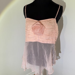 May include: A light pink camisole top with spaghetti straps. The top features a ruched bodice with a large, fabric rose in the center. The bottom portion is a sheer, flowing fabric in a similar shade of pink.