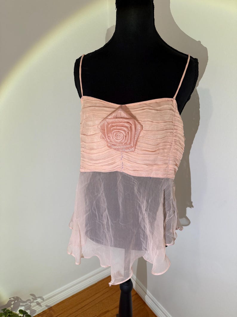 May include: A light pink camisole top with spaghetti straps. The top features a ruched bodice with a large, fabric rose in the center. The bottom portion is a sheer, flowing fabric in a similar shade of pink.