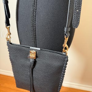 May include: Black crossbody bag with a textured finish and gold-tone hardware. The bag features a long, adjustable strap and a decorative tassel. The edges are stitched with a contrasting thread.
