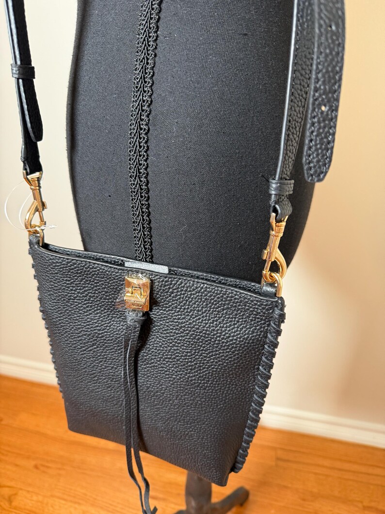 May include: Black crossbody bag with a textured finish and gold-tone hardware. The bag features a long, adjustable strap and a decorative tassel. The edges are stitched with a contrasting thread.