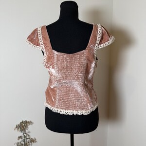 Puede incluir: Top de terciopelo rosa empolvado con cuello cuadrado y mangas cortas. La parte superior presenta un corpi&ntilde;o fruncido y est&aacute; adornada con encaje blanco a lo largo del escote, las mangas y el dobladillo. La prenda se exhibe en un maniqu&iacute; negro.