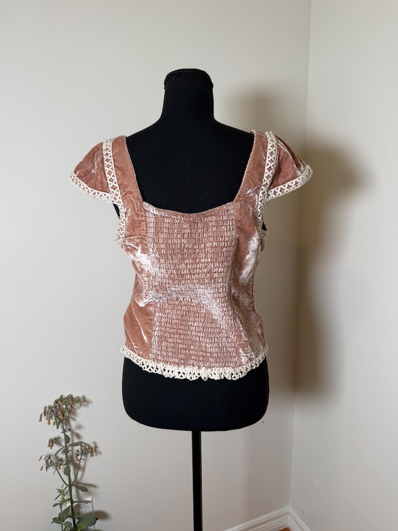 Puede incluir: Top de terciopelo rosa empolvado con cuello cuadrado y mangas cortas. La parte superior presenta un corpi&ntilde;o fruncido y est&aacute; adornada con encaje blanco a lo largo del escote, las mangas y el dobladillo. La prenda se exhibe en un maniqu&iacute; negro.