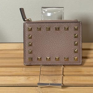 May include: A blush-colored cardholder with gold-toned square studs arranged around the edges. The wallet has a zipper closure and a small leather pull tab. The cardholder is displayed on a clear acrylic stand.