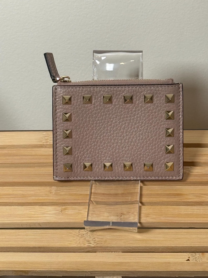 May include: A blush-colored cardholder with gold-toned square studs arranged around the edges. The wallet has a zipper closure and a small leather pull tab. The cardholder is displayed on a clear acrylic stand.