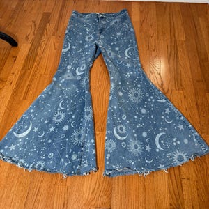 May include: Light blue denim bell-bottom jeans with a celestial print. The design features white suns, moons, and stars scattered across the fabric. The jeans have a classic five-pocket design and a frayed hem.