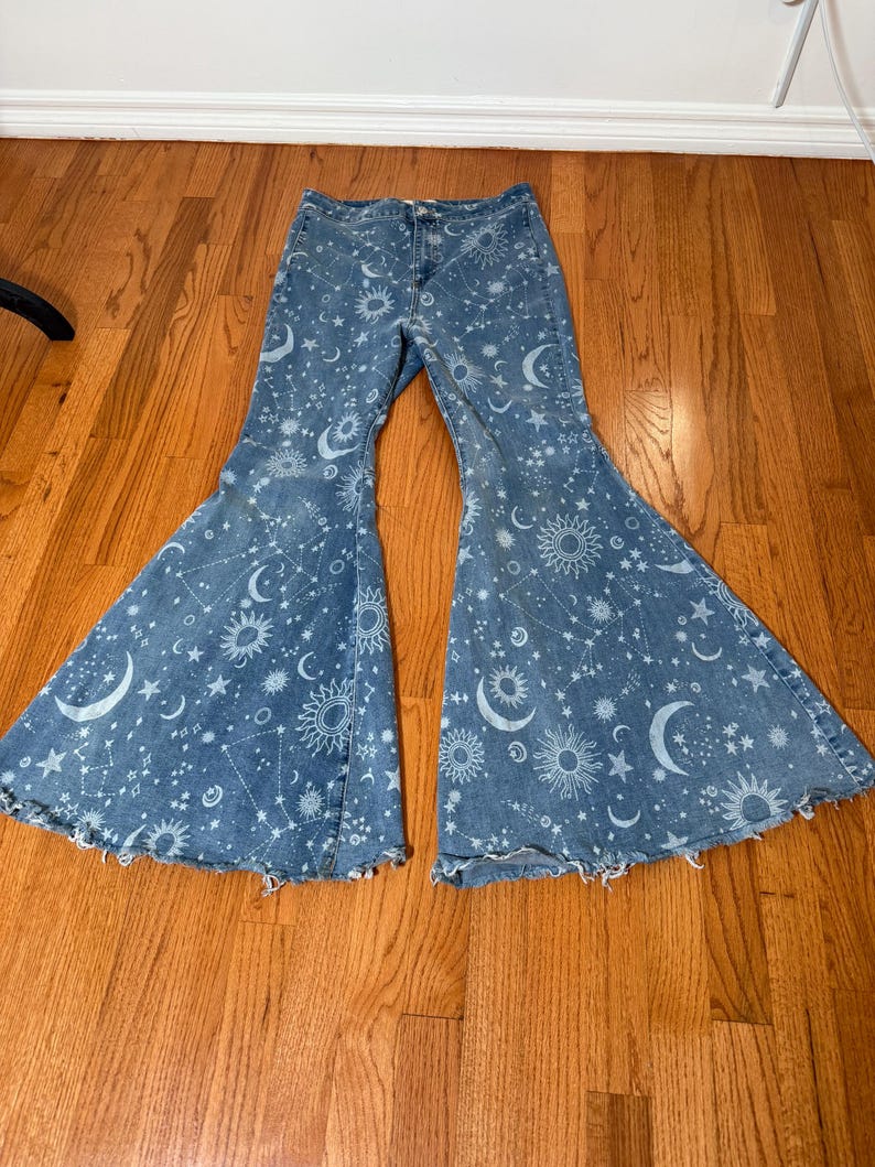 May include: Light blue denim bell-bottom jeans with a celestial print. The design features white suns, moons, and stars scattered across the fabric. The jeans have a classic five-pocket design and a frayed hem.