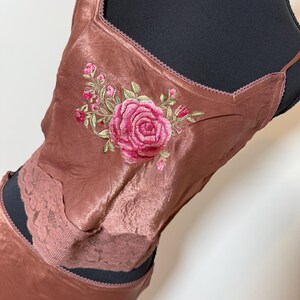 Puede incluir: Conjunto de camisola y falda de sat&eacute;n color rosa empolvado. La camisola presenta un escote cuadrado y est&aacute; adornada con una gran rosa rosa bordada y hojas verdes. La falda tiene un detalle de encaje.