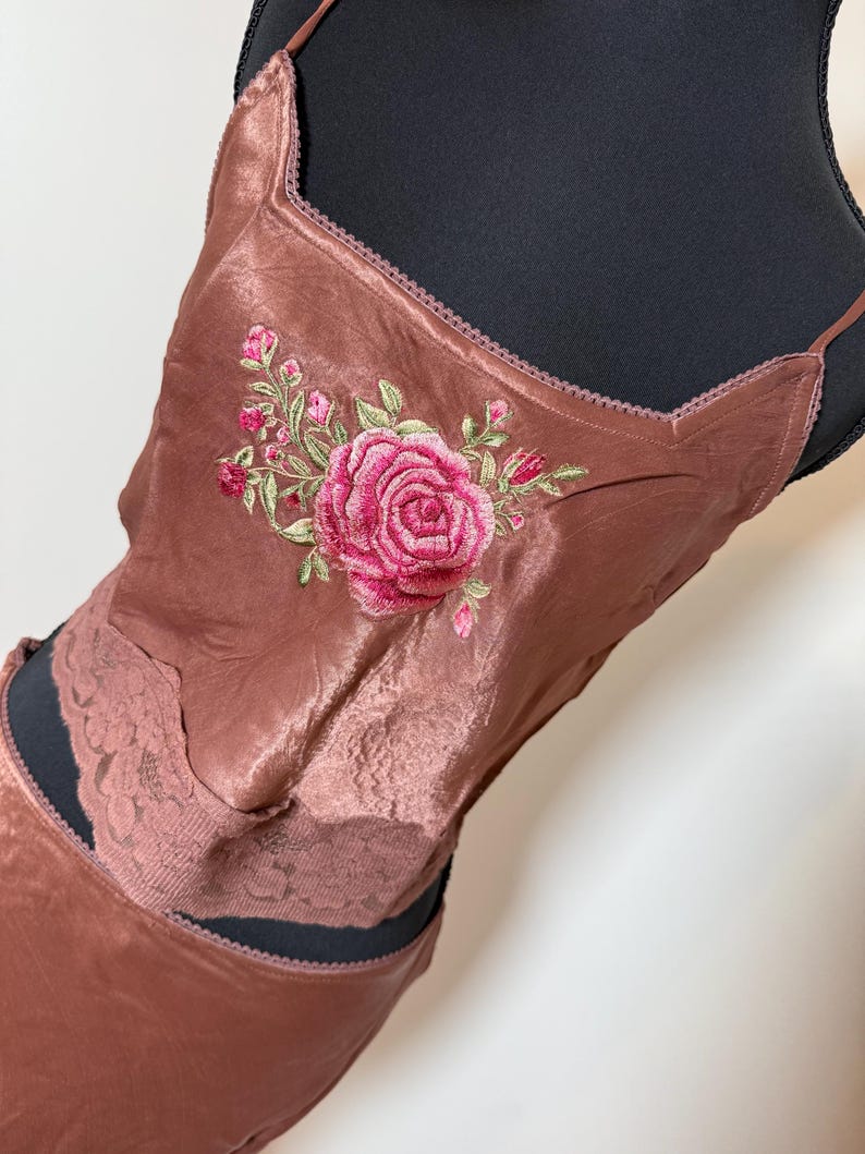 Puede incluir: Conjunto de camisola y falda de sat&eacute;n color rosa empolvado. La camisola presenta un escote cuadrado y est&aacute; adornada con una gran rosa rosa bordada y hojas verdes. La falda tiene un detalle de encaje.