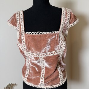 Puede incluir: Top de terciopelo rosa empolvado con cuello cuadrado y mangas cortas. El top presenta un ribete de encaje blanco a lo largo del escote, las mangas y las costuras del corpi&ntilde;o. La prenda se exhibe en un maniqu&iacute; negro.