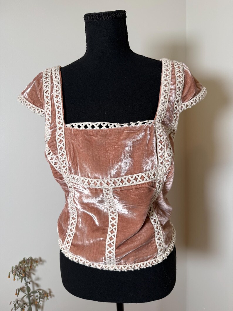 Puede incluir: Top de terciopelo rosa empolvado con cuello cuadrado y mangas cortas. El top presenta un ribete de encaje blanco a lo largo del escote, las mangas y las costuras del corpi&ntilde;o. La prenda se exhibe en un maniqu&iacute; negro.