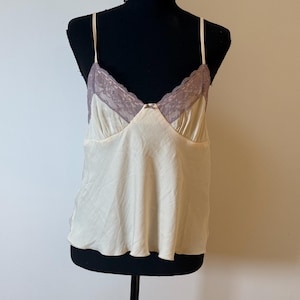 May include: Cream-colored camisole with thin straps and a lace trim in a mauve hue. The top is displayed on a black mannequin, showcasing its delicate design and silky texture. A small bow detail is visible at the neckline.