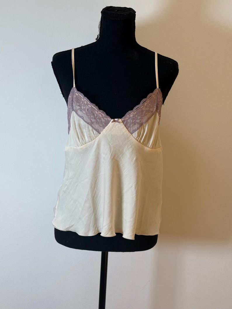 May include: Cream-colored camisole with thin straps and a lace trim in a mauve hue. The top is displayed on a black mannequin, showcasing its delicate design and silky texture. A small bow detail is visible at the neckline.