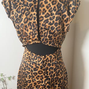 Puede incluir: Un conjunto de dos piezas con estampado de leopardo. La parte superior tiene mangas cortas y una espalda con abertura. La falda tiene cintura alta y una fila de botones en la espalda. La tela es un estampado de animales marr&oacute;n y negro.