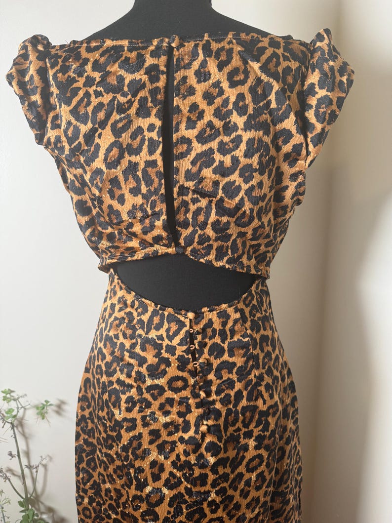 Puede incluir: Un conjunto de dos piezas con estampado de leopardo. La parte superior tiene mangas cortas y una espalda con abertura. La falda tiene cintura alta y una fila de botones en la espalda. La tela es un estampado de animales marr&oacute;n y negro.