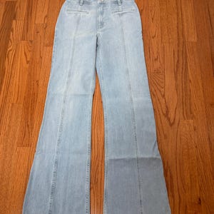 Puede incluir: Pantalones vaqueros azul claro con cintura alta y perneras acampanadas. Los vaqueros tienen un dise&ntilde;o cl&aacute;sico de cinco bolsillos y costuras verticales en la parte delantera y trasera de las perneras. Los pantalones est&aacute;n sobre una superficie de madera.
