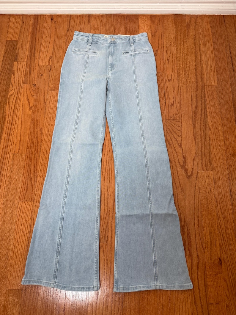 Puede incluir: Pantalones vaqueros azul claro con cintura alta y perneras acampanadas. Los vaqueros tienen un dise&ntilde;o cl&aacute;sico de cinco bolsillos y costuras verticales en la parte delantera y trasera de las perneras. Los pantalones est&aacute;n sobre una superficie de madera.
