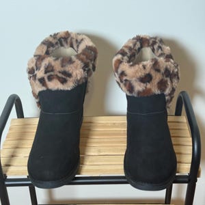 Puede incluir: Un par de botas de ante negro con ribete de estampado de leopardo. Las botas se exhiben en una estantería de metal negro y bambú contra una pared blanca. Las botas tienen punta redonda y tacón bajo.