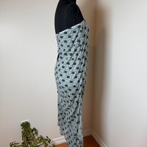 May include: A pale blue dress with a black floral design is displayed on a black mannequin. The dress is draped and has a unique texture. The dress is sleeveless and appears to be made of a lightweight fabric.