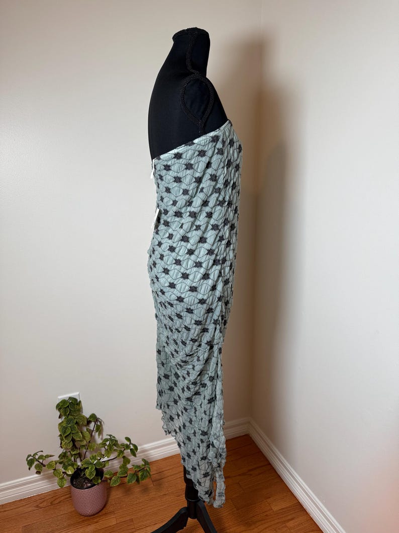 May include: A pale blue dress with a black floral design is displayed on a black mannequin. The dress is draped and has a unique texture. The dress is sleeveless and appears to be made of a lightweight fabric.