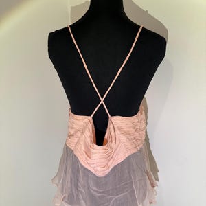 May include: A light pink camisole top with spaghetti straps that cross in the back. The top features a ruched bodice and a flowing, sheer skirt. The garment is displayed on a black mannequin.