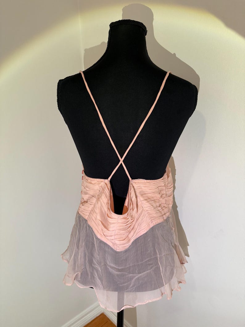 May include: A light pink camisole top with spaghetti straps that cross in the back. The top features a ruched bodice and a flowing, sheer skirt. The garment is displayed on a black mannequin.