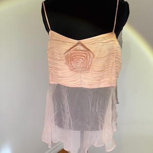 May include: A light pink camisole top with spaghetti straps. The top features a pleated bodice with a rose applique and a sheer, ruffled hem. The garment is displayed on a black mannequin.