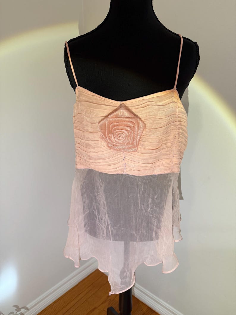 May include: A light pink camisole top with spaghetti straps. The top features a pleated bodice with a rose applique and a sheer, ruffled hem. The garment is displayed on a black mannequin.