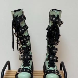 May include: A pair of mint green knee-high boots featuring black laces and multiple black buckled straps with silver-toned metal eyelets. The boots have a thick, chunky black platform sole and a side zipper closure for a secure fit.