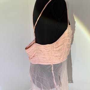 May include: A light pink top with thin straps and a gathered bodice. The top is paired with a sheer, flowing skirt. The garment is displayed on a black mannequin, showcasing its design and texture.