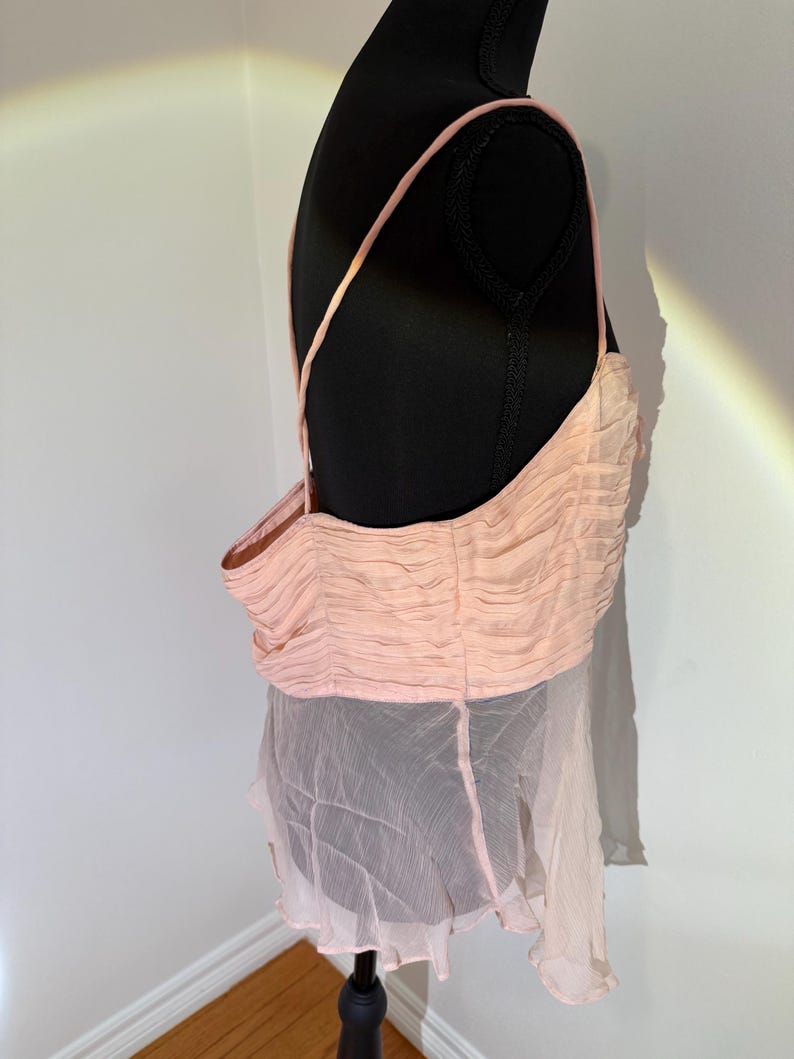 May include: A light pink top with thin straps and a gathered bodice. The top is paired with a sheer, flowing skirt. The garment is displayed on a black mannequin, showcasing its design and texture.