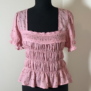 May include: A light pink top with a square neckline and short, ruffled sleeves. The top features a smocked bodice with a ruffled hem and is adorned with small, dark brown floral patterns. The garment is displayed on a black mannequin.