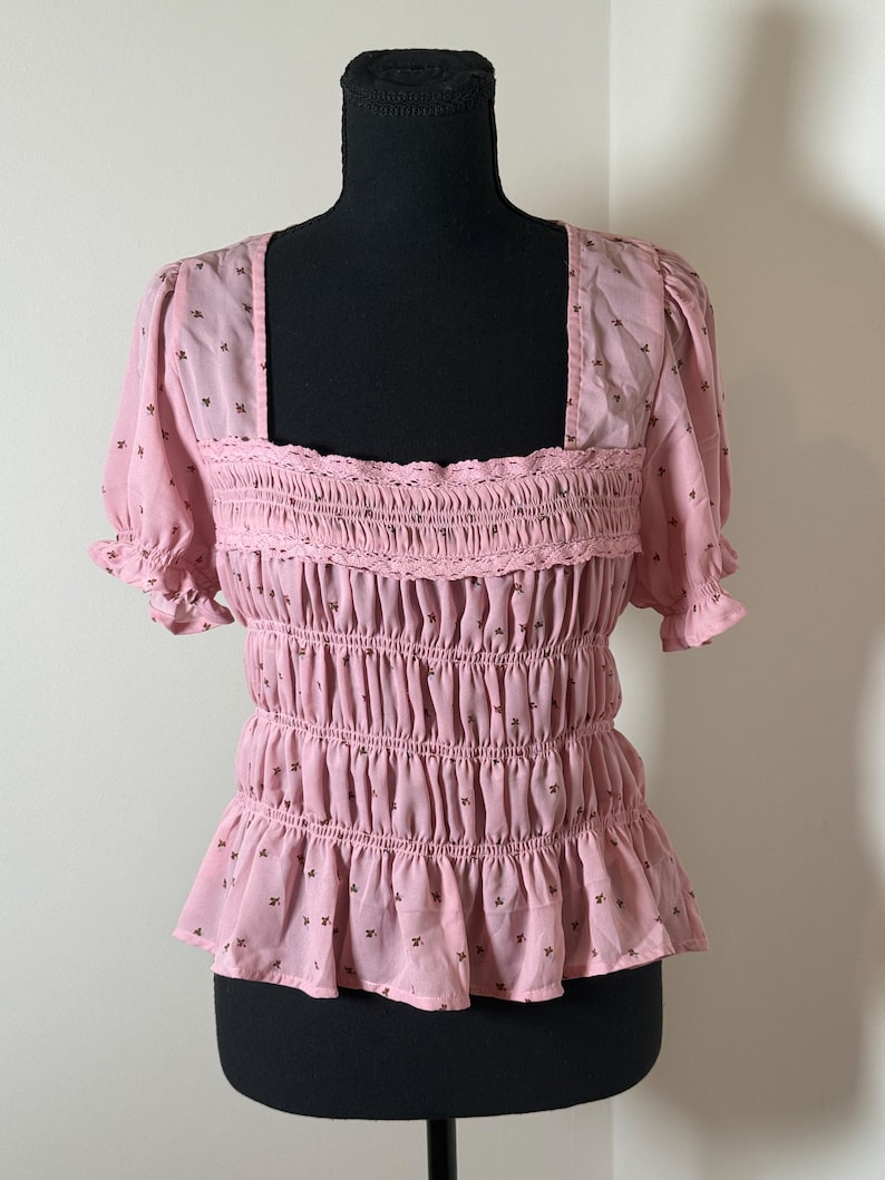 May include: A light pink top with a square neckline and short, ruffled sleeves. The top features a smocked bodice with a ruffled hem and is adorned with small, dark brown floral patterns. The garment is displayed on a black mannequin.