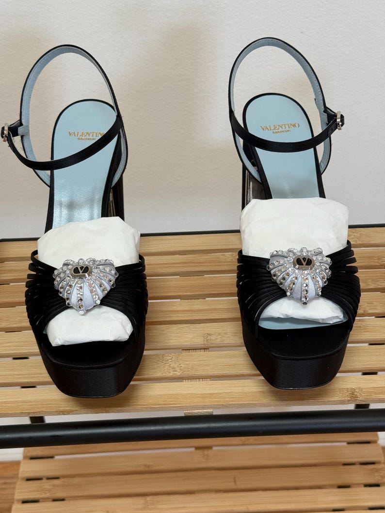 May include: A pair of black satin platform sandals with ankle straps and a light blue insole featuring the Valentino Garavani logo. The front of each shoe is adorned with a heart-shaped embellishment covered in clear crystals and a central V logo.