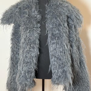 May include: A gray, shaggy faux fur jacket displayed on a black mannequin. The jacket has long, textured fibers, a collar, and open front. The jacket is a neutral color and appears soft and warm.
