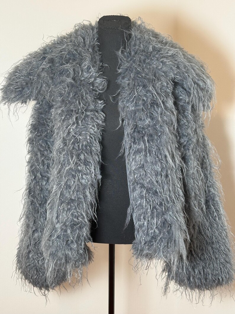 May include: A gray, shaggy faux fur jacket displayed on a black mannequin. The jacket has long, textured fibers, a collar, and open front. The jacket is a neutral color and appears soft and warm.
