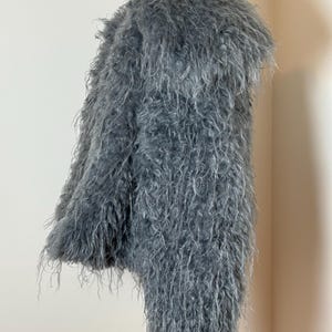 May include: A gray, shaggy coat displayed on a mannequin. The coat has a long, textured pile, giving it a fluffy appearance. The garment is a solid gray color and appears to be made of a soft material.