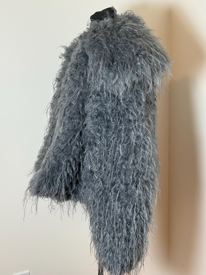 May include: A gray, shaggy coat displayed on a mannequin. The coat has a long, textured pile, giving it a fluffy appearance. The garment is a solid gray color and appears to be made of a soft material.