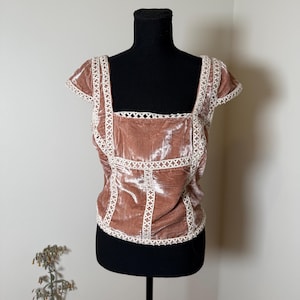 Puede incluir: Top de terciopelo rosa empolvado con cuello cuadrado y mangas cortas. El top est&aacute; adornado con detalles de encaje blanco a lo largo del escote, las mangas y las costuras. La prenda se exhibe en un maniqu&iacute; negro.