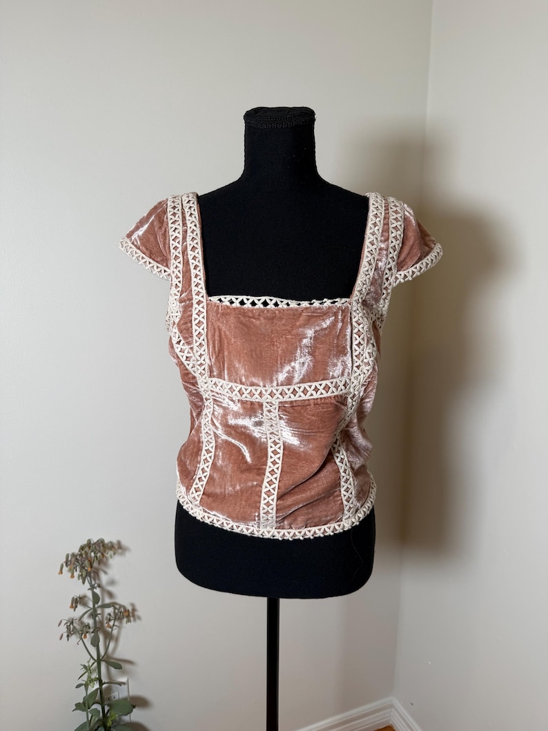 Puede incluir: Top de terciopelo rosa empolvado con cuello cuadrado y mangas cortas. El top est&aacute; adornado con detalles de encaje blanco a lo largo del escote, las mangas y las costuras. La prenda se exhibe en un maniqu&iacute; negro.