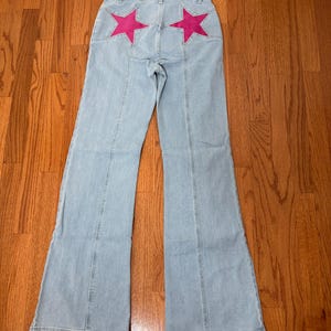 Puede incluir: Vaqueros de mezclilla azul claro con dise&ntilde;o de pierna acampanada. Los bolsillos traseros est&aacute;n adornados con grandes aplicaciones de estrellas rosas brillantes. Los vaqueros tienen un dise&ntilde;o cl&aacute;sico de cinco bolsillos y cierre de bot&oacute;n y cremallera.