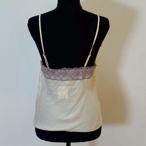 May include: Ivory-colored camisole with thin straps and a lavender lace trim. The top is displayed on a black mannequin. The fabric appears to be a lightweight, silky material, suitable for layering or sleepwear.