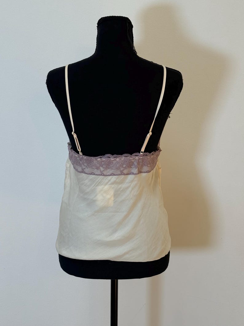 May include: Ivory-colored camisole with thin straps and a lavender lace trim. The top is displayed on a black mannequin. The fabric appears to be a lightweight, silky material, suitable for layering or sleepwear.