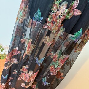 以下が含まれることがあります： 赤、ピンク、緑、青の色合いで刺繍された花柄と蝶柄が施された黒のシアー生地。生地は重ねられており、半透明の効果を生み出しています。刺繍は衣服に質感と立体感を与えています。
