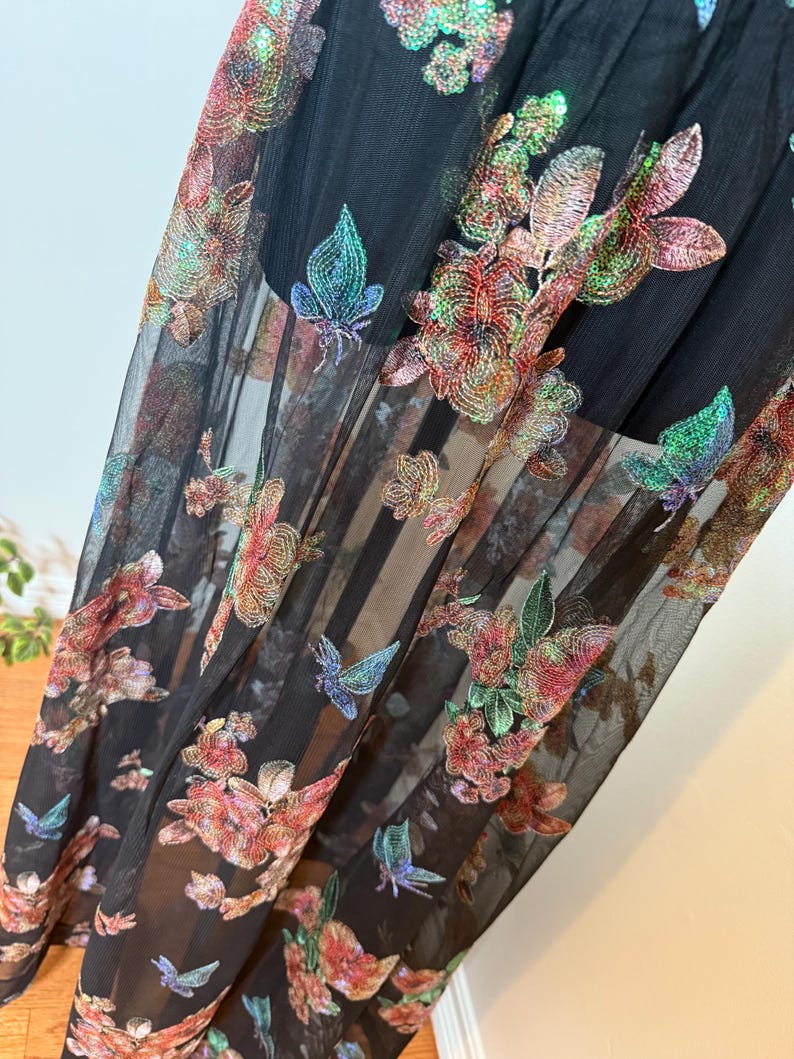 以下が含まれることがあります： 赤、ピンク、緑、青の色合いで刺繍された花柄と蝶柄が施された黒のシアー生地。生地は重ねられており、半透明の効果を生み出しています。刺繍は衣服に質感と立体感を与えています。
