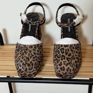 May include: A pair of platform sandals with a leopard print pattern. The sandals have a thick sole, an ankle strap, and a rounded toe. The brand name "KELSI DAGGER" is visible on the insole. The sandals are displayed on a wooden shelf.